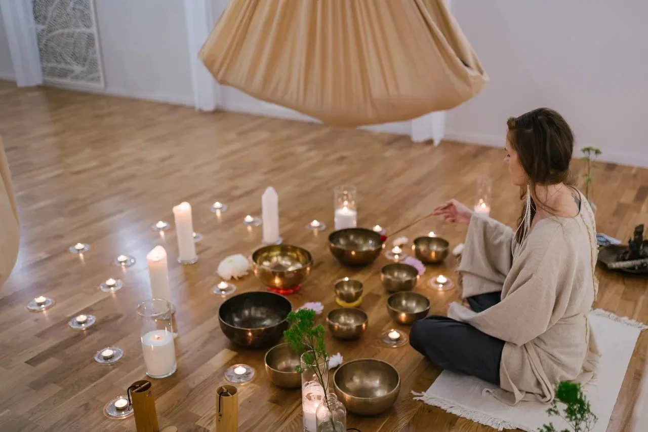 Женщина в бежевом халате сидит на полу среди поющих чаш и свечей в спокойном помещении