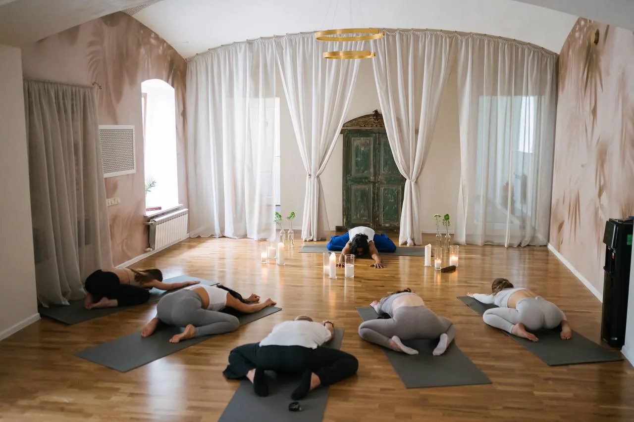 Групповое занятие по йоге и медитации в студии, люди в позе ребёнка на ковриках вокруг небольшого алтаря с свечами и растениями