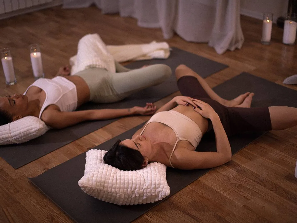 Две женщины лежат на ковриках для йоги в затемнённом зале с свечами, расслабляясь на валиках во время практики отдыха